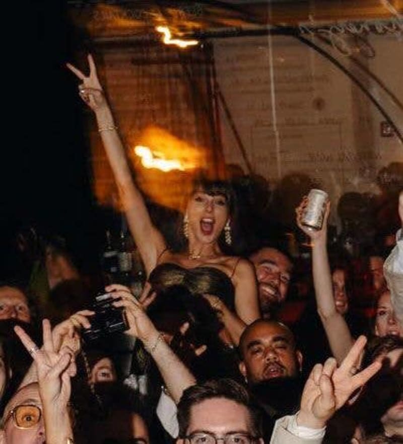 A woman in a black dress celebrates with raised arm among a crowd of people at an indoor party or nightclub event.