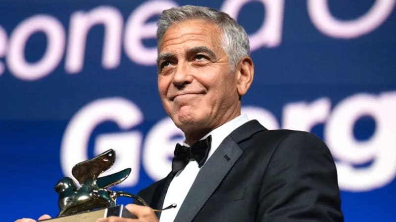 Man in black tuxedo holding winged lion trophy, smiling, at Venice Film Festival ceremony.