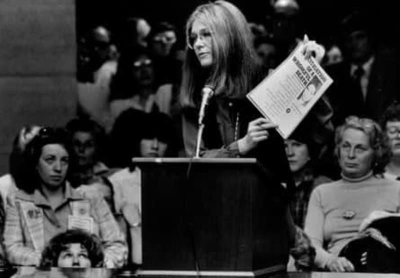 Woman with long hair speaking at podium holding newspaper at what appears to be a 1970s public event or rally.