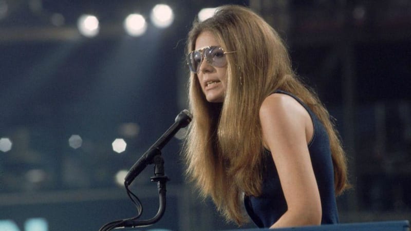 A woman with long brown hair and aviator sunglasses performs at a microphone on stage with stage lighting in the background.
