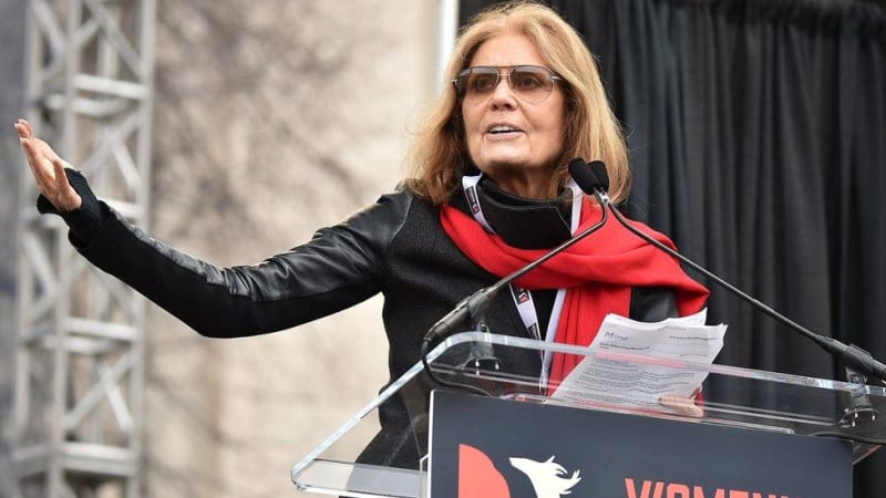 A person with long blonde hair wearing sunglasses, a black leather jacket, and red scarf speaks at a podium with papers.