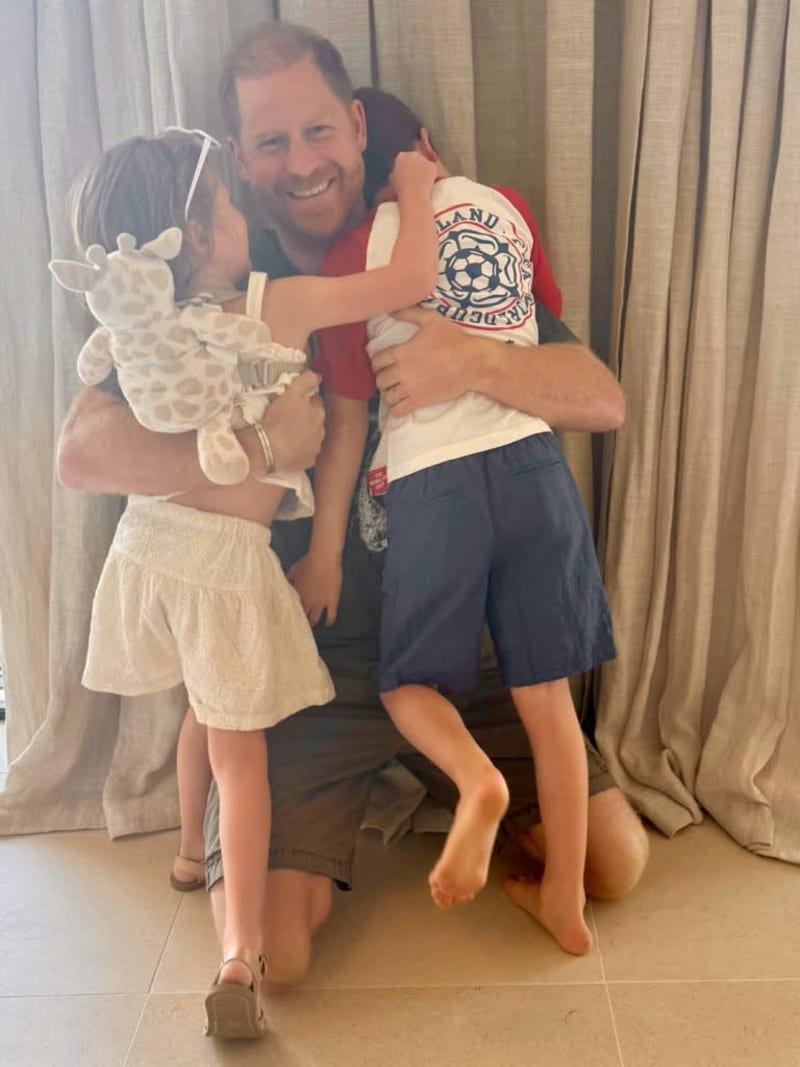 A man in a red shirt smiles while holding two young children, one carrying a stuffed animal, in front of beige curtains.