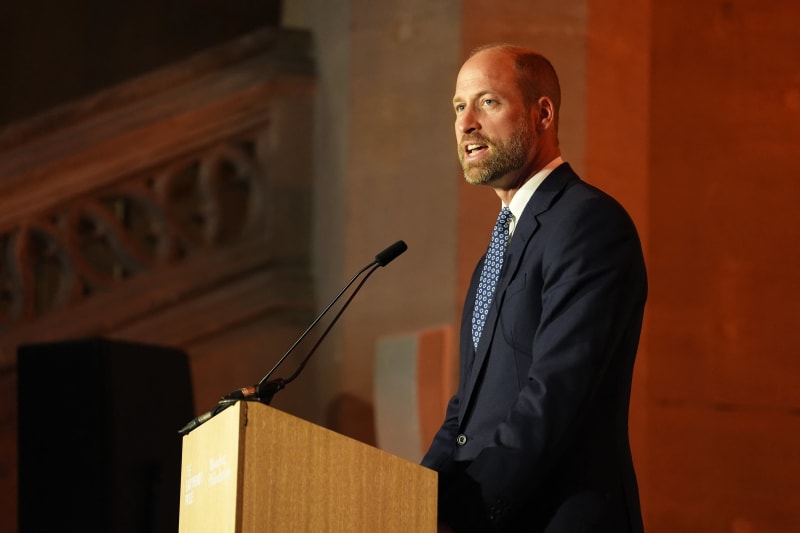 A man in a dark suit and blue tie speaks at a wooden podium with a microphone in an auditorium with orange walls.