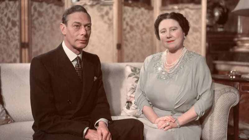 Two people in formal attire sitting on a cream-colored sofa in an elegant interior setting with patterned wallpaper.