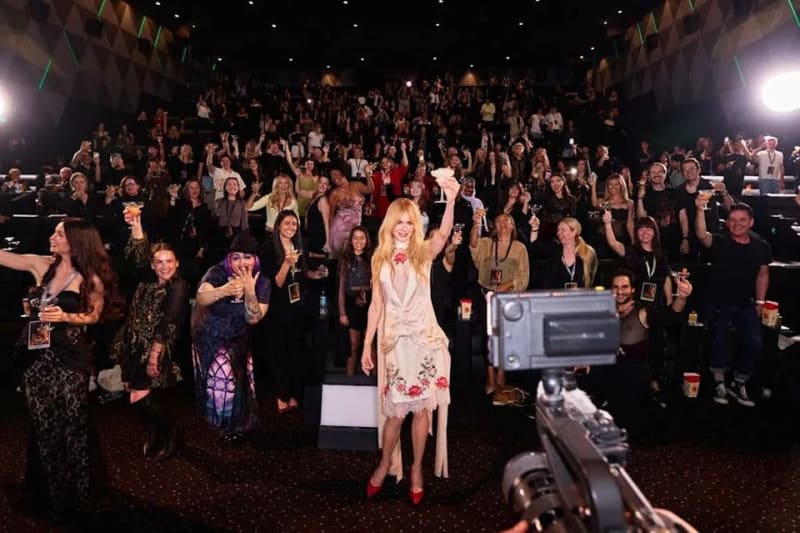 A blonde woman in a floral dress celebrates among a crowd of people holding drinks at what appears to be an indoor event or party.