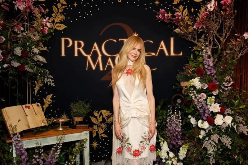 Woman in white dress with floral embroidery at Practical Magic premiere event with decorated backdrop and flowers.