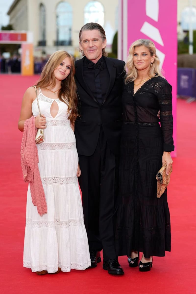 Three people pose together on a red carpet at an outdoor event, with a pink and white building visible in the background.