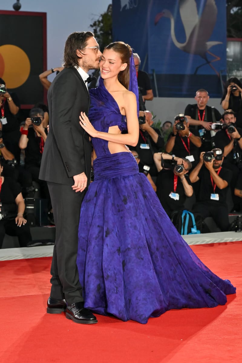 Man in black suit kissing woman's cheek, woman in purple one-shoulder gown, red carpet with photographers behind.