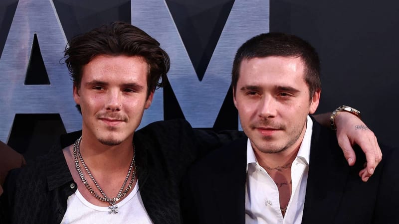 Two men in formal black attire pose together at an event with large illuminated letters visible in the background.