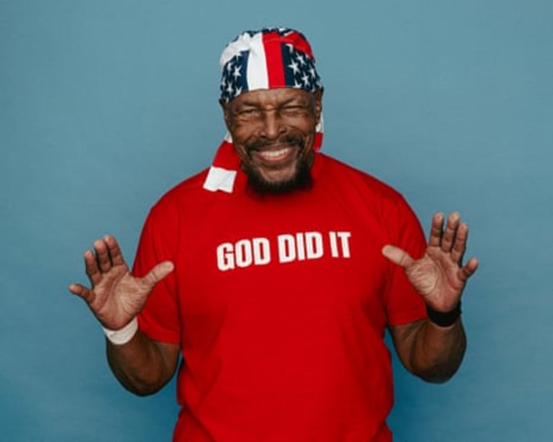 A man wearing a red "God Did It" shirt and patriotic headwrap smiles with hands raised against a blue background.