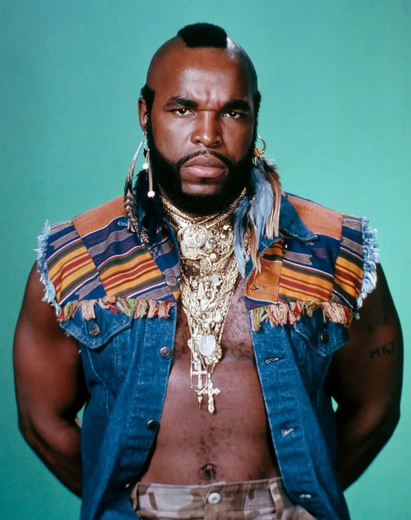Man in denim vest with gold chains and mohawk, studio portrait against teal background.