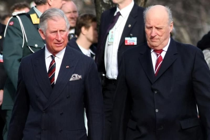 Two men in formal dark suits with red ties walk together at an official ceremony, surrounded by military and dignitaries.