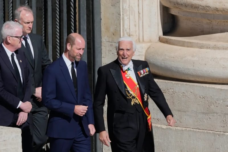 Man in navy suit walks alongside elderly man in black tails with red sash and medals, stone building exterior.