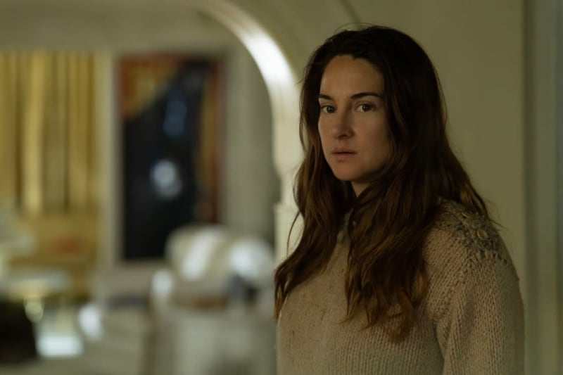 A woman with long dark hair wearing a beige sweater looks over her shoulder in an interior hallway setting.