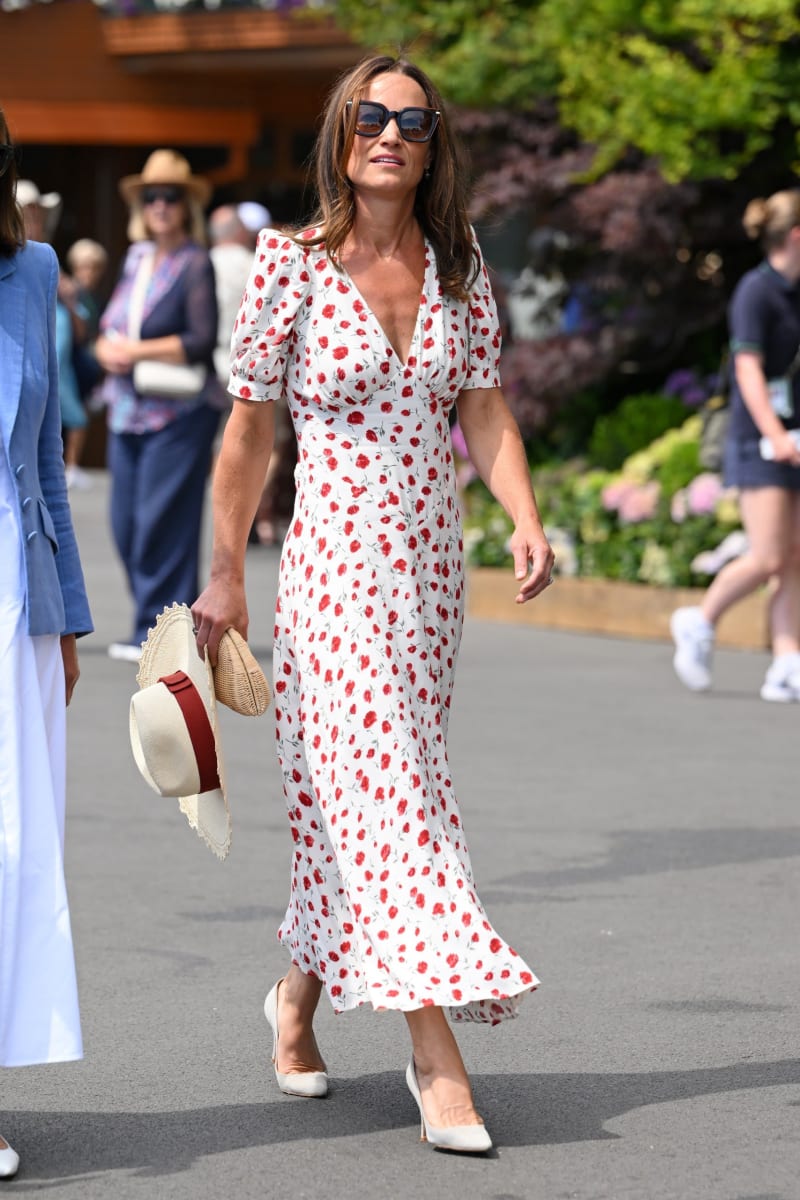 Woman in white red floral midi dress, sunglasses, carrying straw hat, walking outdoors.