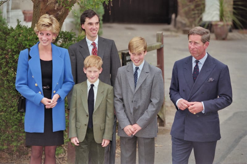 Five people in formal attire stand together outdoors in front of ivy-covered wall and courtyard setting.
