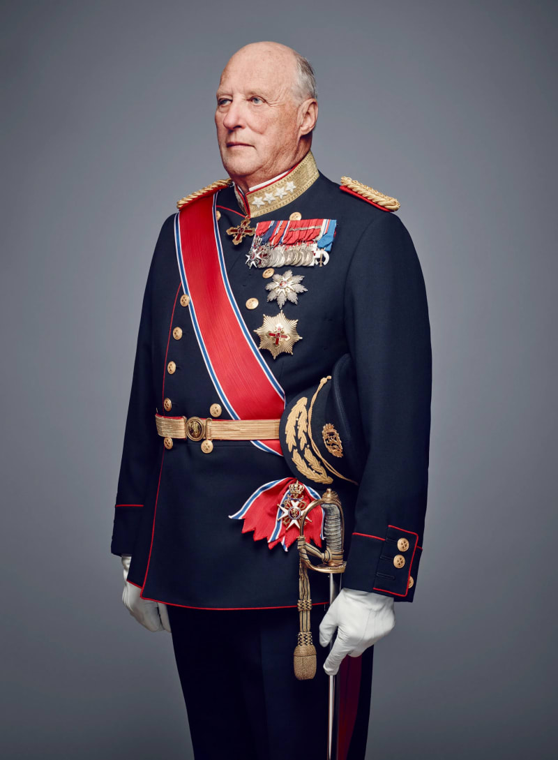 Elderly man in navy military uniform with red sash, medals, and ceremonial sword, standing against grey background.