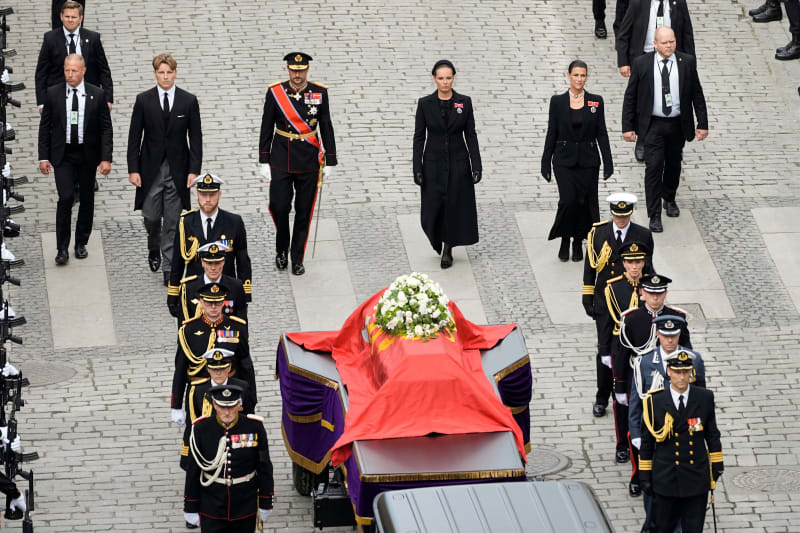 A formal state funeral procession with military personnel in dress uniforms escorting a draped coffin through a brick courtyard.
