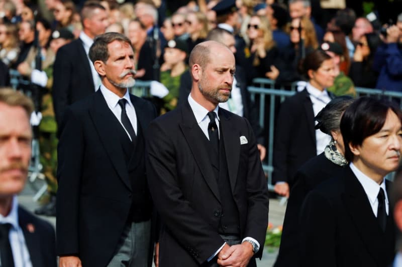 Dignitaries in formal black attire walk solemnly among a crowd at what appears to be an official state or funeral ceremony.
