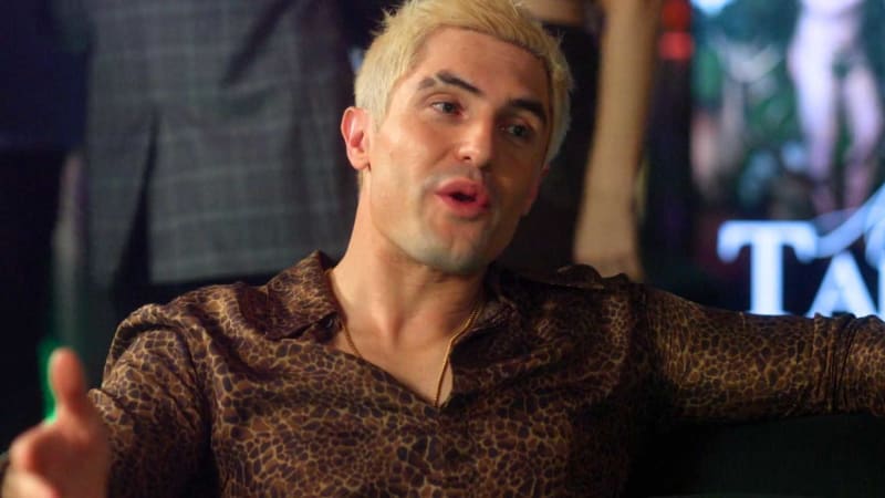 A man with blonde hair wearing a leopard print shirt speaks expressively during what appears to be a televised interview or talk show.