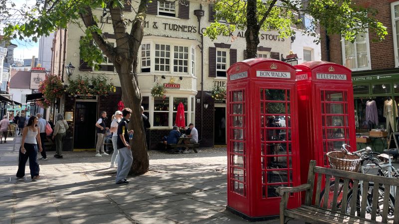 Red British telephone boxes in a sunny London street plaza with pedestrians and historic buildings.