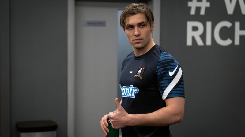 Man in navy and blue Nike football shirt with 'bantr' sponsor, holding bottle, indoor stadium corridor.