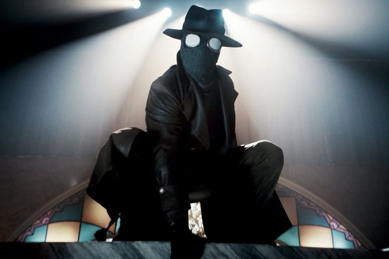 Figure in black trench coat, mask, goggles and fedora, seated under spotlights in ornate interior.