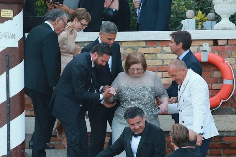 A woman in a gray dress is assisted by several men in suits at what appears to be an official outdoor ceremony near a brick building.