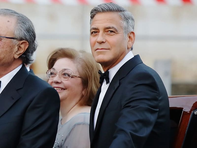 Man in black tuxedo seated beside woman in grey top with glasses at outdoor event.