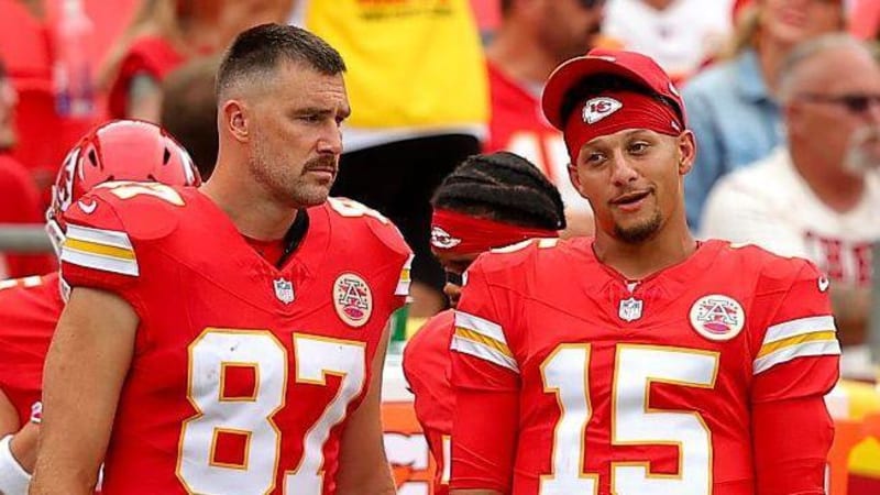 Three Kansas City Chiefs players in red uniforms stand together on the sideline during a game.