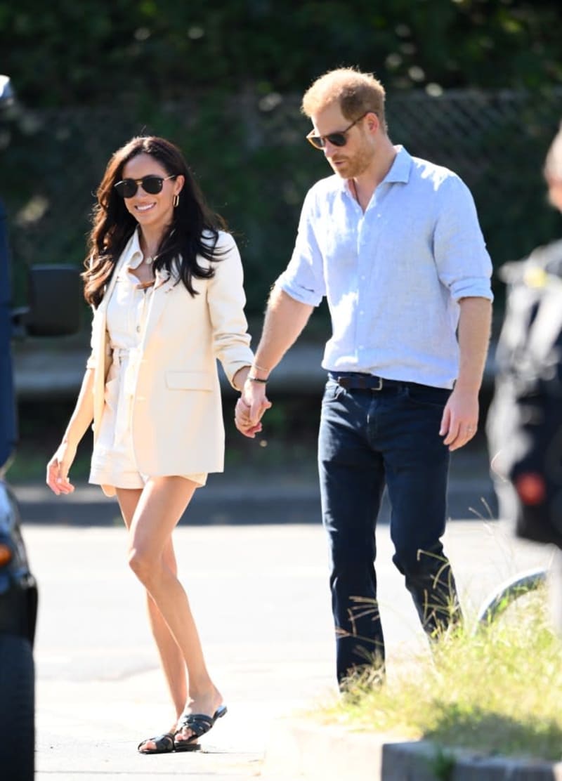 A couple walks hand-in-hand outdoors, the woman wearing a cream blazer and sunglasses, the man in a light blue shirt and dark jeans.