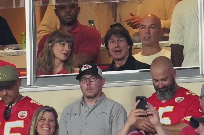 People watch a Kansas City Chiefs game from a luxury box, with spectators in red and gold team apparel visible both inside and outside the window.