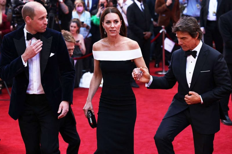 Two men in tuxedos and woman in black off-shoulder gown walking red carpet.