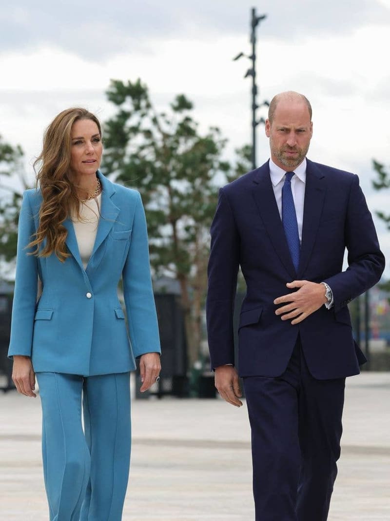 Two people in formal business attire walking outdoors on a paved area with trees in the background under cloudy skies.