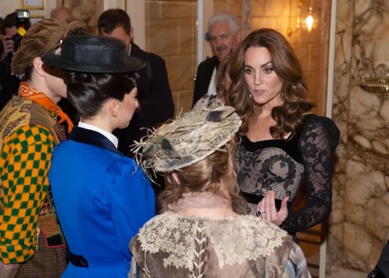 A woman in a black lace dress converses with elegantly dressed attendees at a formal indoor event.