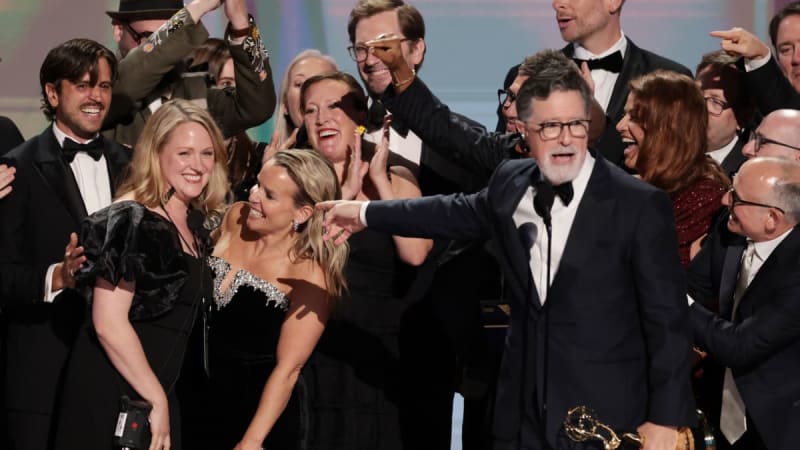 A group of formally dressed people celebrate on stage at an awards ceremony, smiling and gesturing enthusiastically together.