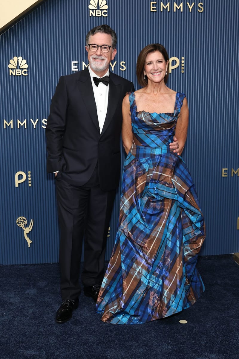 Two people at the Emmy Awards red carpet, man in black tuxedo with glasses, woman in blue plaid dress.