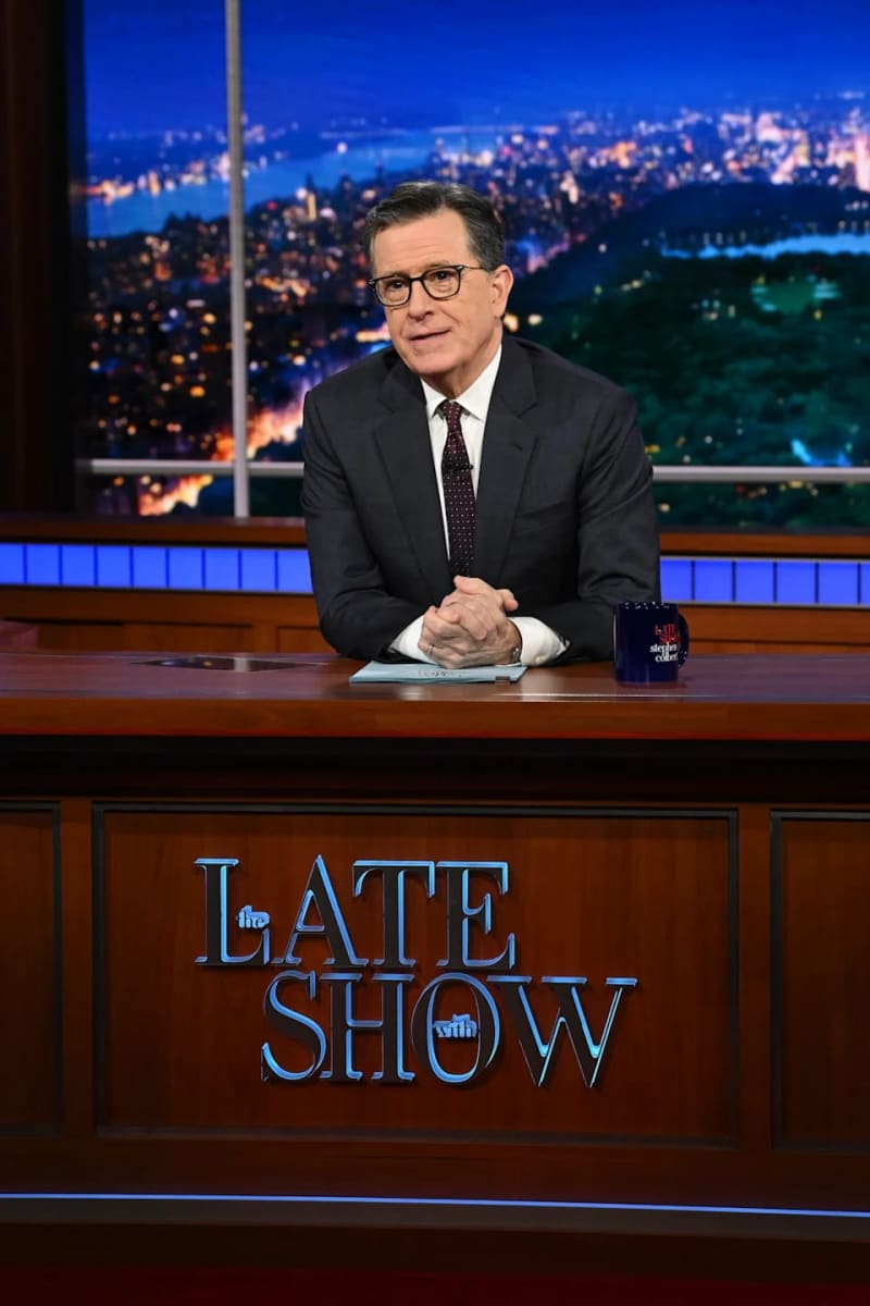 A man in a dark suit and glasses sits at a desk on the set of The Late Show with a cityscape visible behind him.