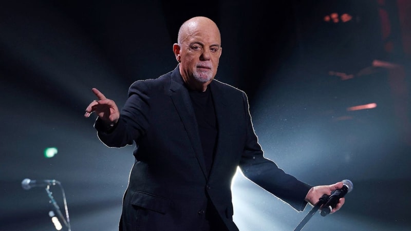 A bald man in a dark blazer holds a microphone while gesturing expressively on stage under dramatic blue lighting.