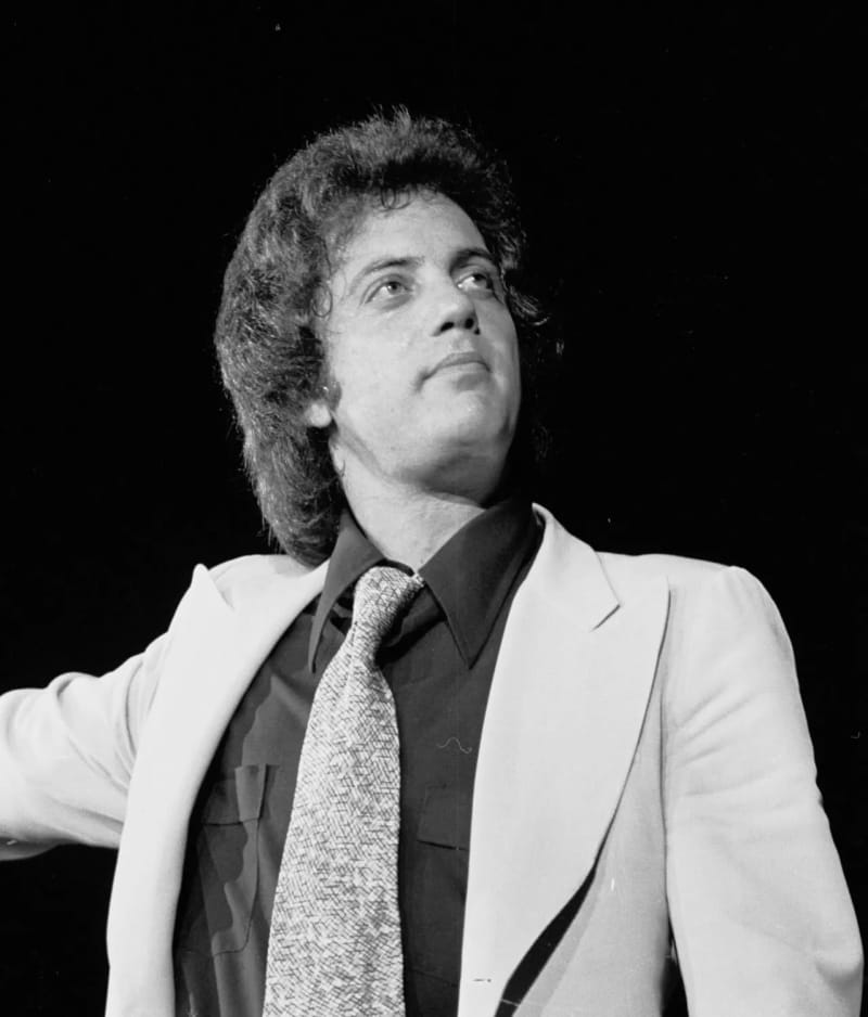 Man in white blazer, dark shirt and tie, looking upward, black background.