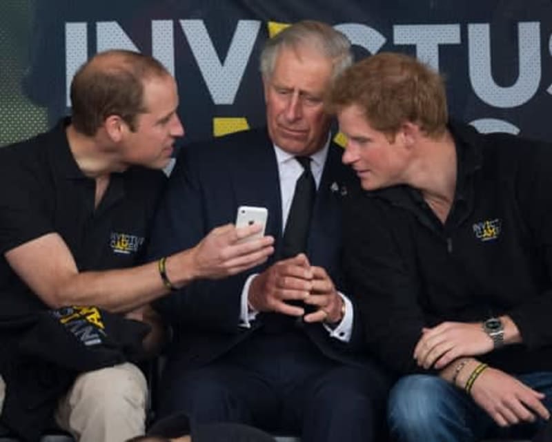 Three men in dark clothing sit together at an event, with one holding a smartphone while they engage in conversation.