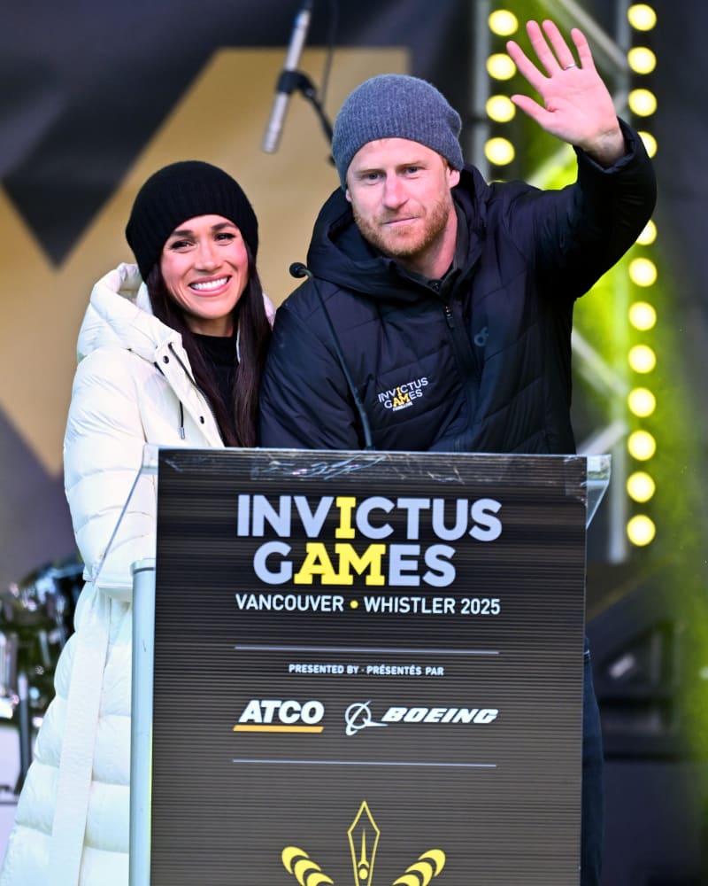 Woman in white puffer coat and man in black jacket waving at Invictus Games Vancouver Whistler 2025 podium.
