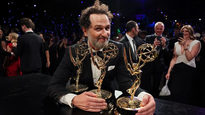 A bearded man in a black tuxedo holds multiple Emmy Awards at an elegant evening event with guests in the background.