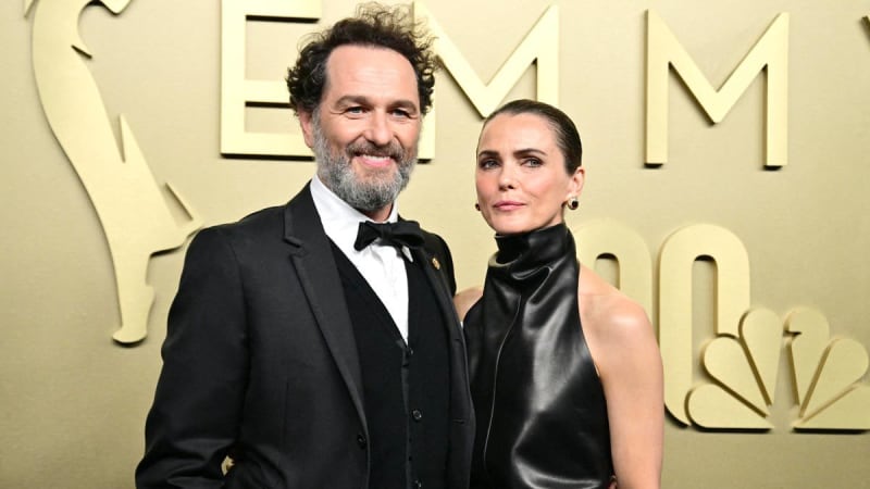 Two people in formal black attire pose together at what appears to be an elegant event with a cream and gold backdrop.