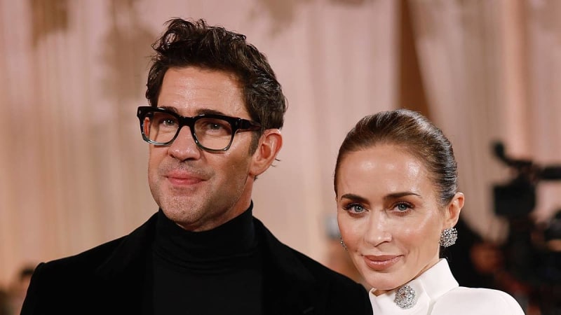 Two people at a formal event; man wearing black turtleneck and glasses, woman in white outfit with diamond jewelry.