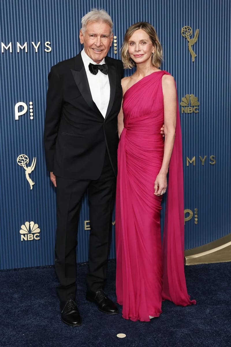 Two people in formal attire pose together at the Emmy Awards, with the man wearing a black tuxedo and the woman in a pink gown.
