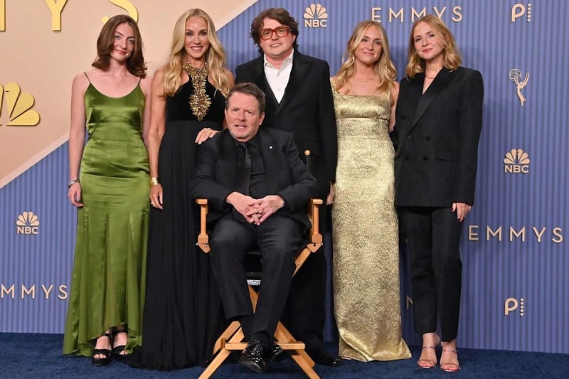 Man seated in director's chair in black suit, surrounded by five family members in green, black and gold formal wear, at Emmy Awards step-and-repeat.