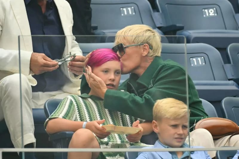 A woman with short blonde hair and sunglasses sits with two young children, one with bright pink hair, in stadium seating.