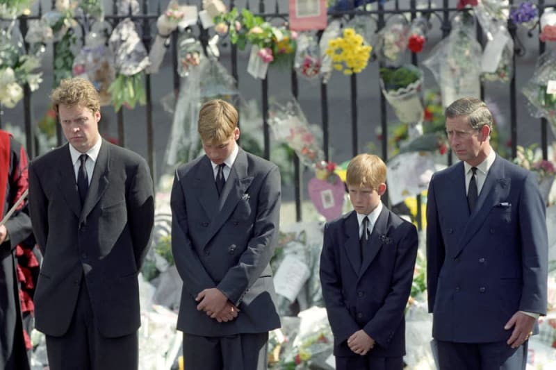 Four males in dark suits standing solemnly at a cemetery with flowers and wreaths visible on the iron fence behind them.