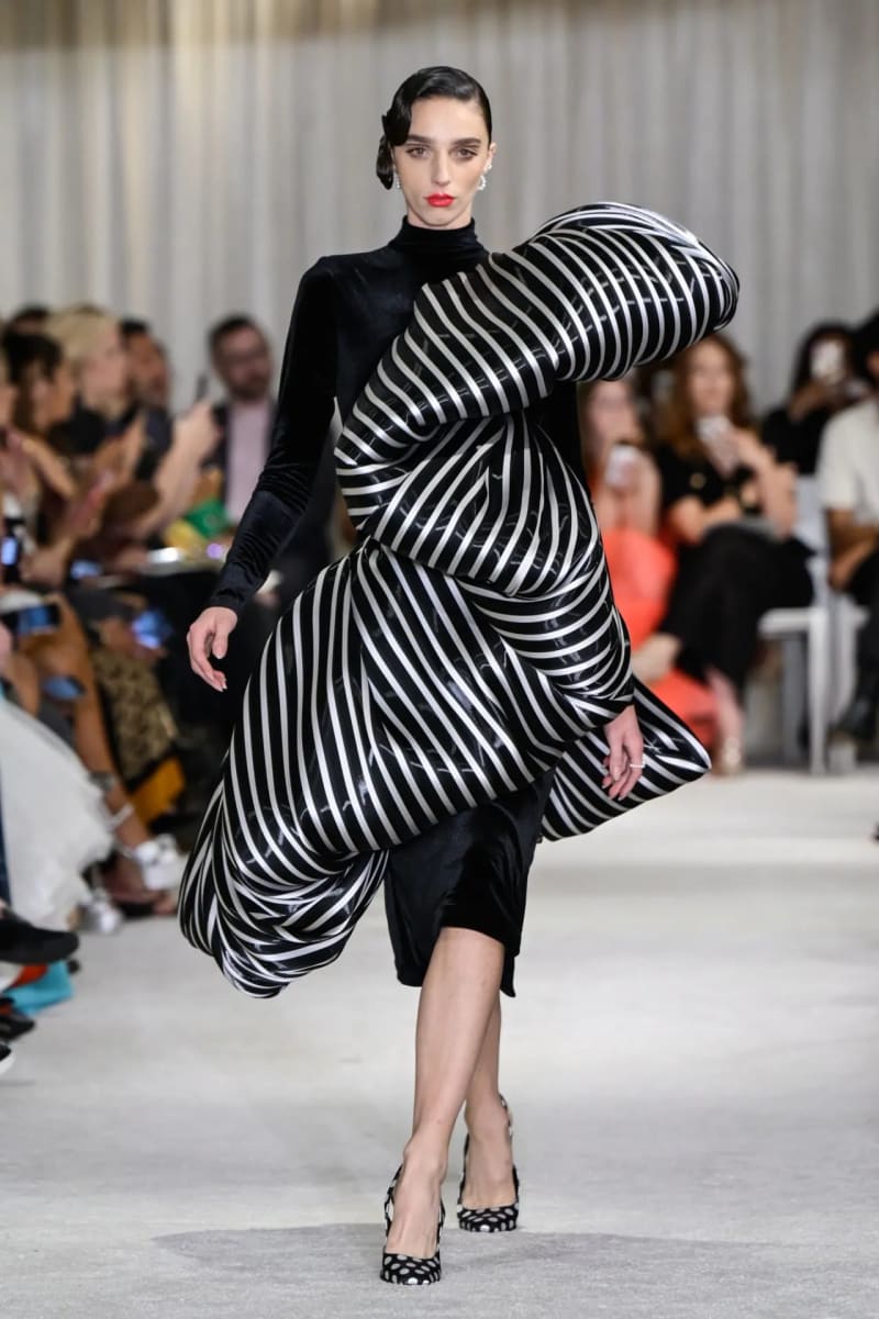 Woman in black velvet dress with oversized black-and-white striped inflated sculptural overlay, walking runway.